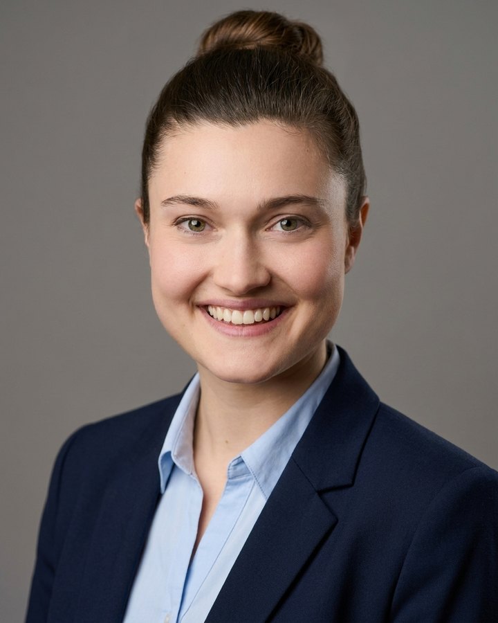 The AI-generated headshot of the same woman: grey studio backdrop, navy blazer, light-blue shirt.